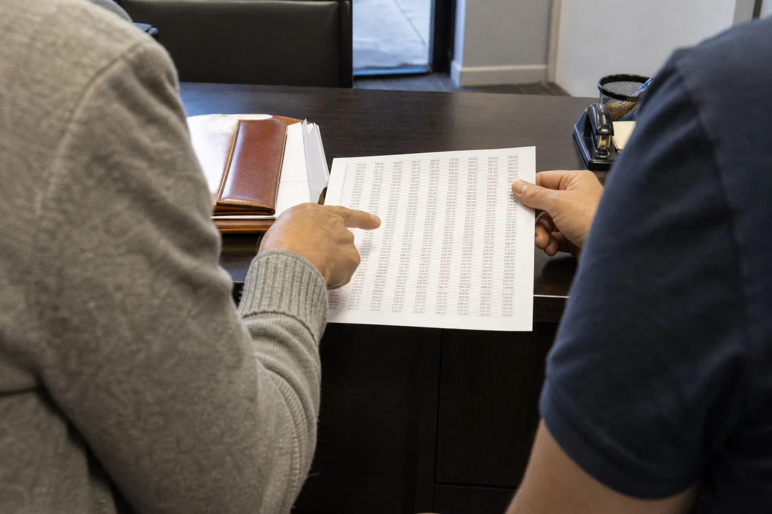 Over-the-shoulder view of two people at a dark office desk reviewing a printed table of figures, one pointing at a column, with a leather portfolio and stapler nearby